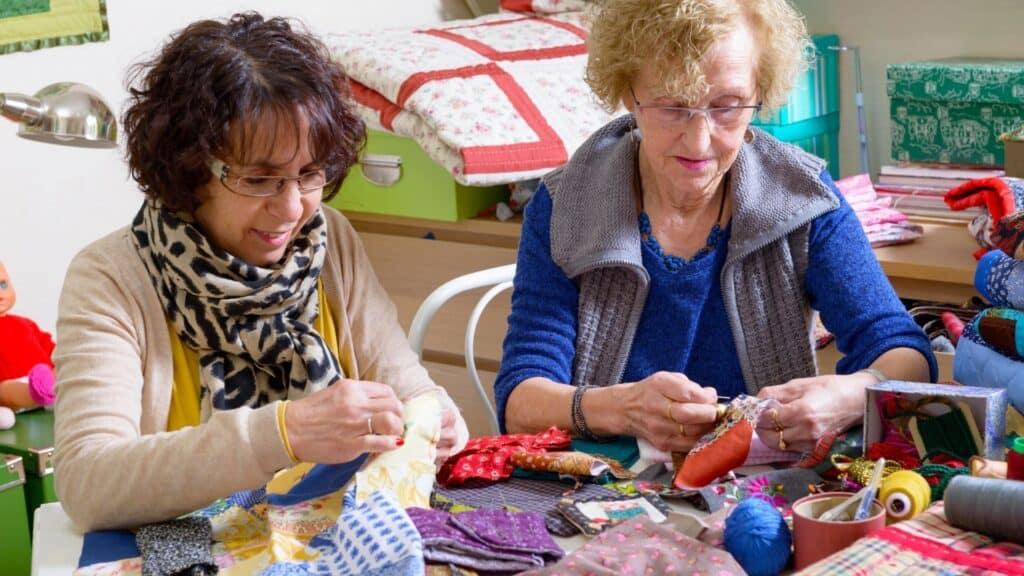 Two women are hand-sewing quilt tops at a table.